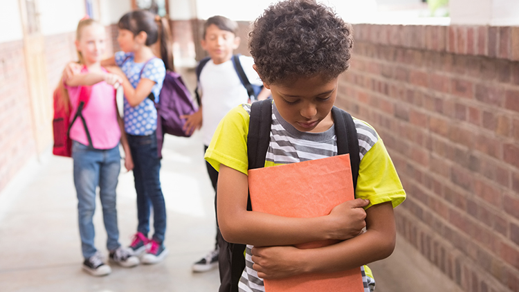 Waist-length selective focus view of a young boy looking sad while being teased by classmates.