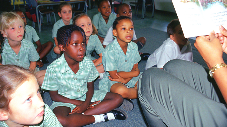 Children in Johannesburgh, South Africa sitting in a carpet as an adult reads a book to them.