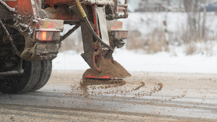 View from the car behind orange highway maintenance truck spreading de-icing salt and sand, crystals dropping on the ice-covered asphalt road.