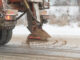 View from the car behind orange highway maintenance truck spreading de-icing salt and sand, crystals dropping on the ice-covered asphalt road.